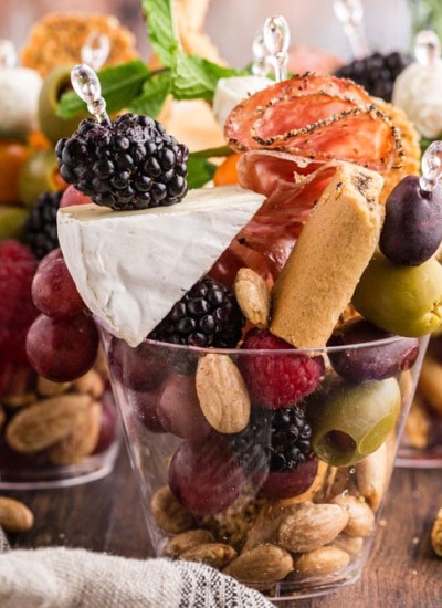 three charcuterie cups filled with assorted snack foods, placed on a wooden surface with additional snacks on a platter in the background and a white cloth beside the cups