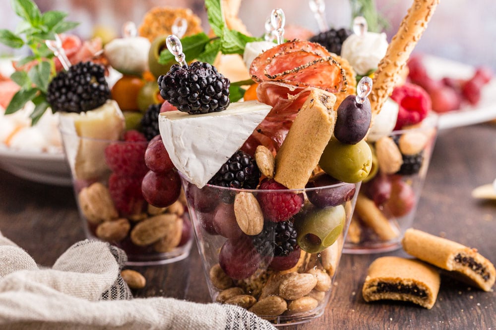 three charcuterie cups filled with assorted snack foods, placed on a wooden surface with additional snacks on a platter in the background and a white cloth beside the cups