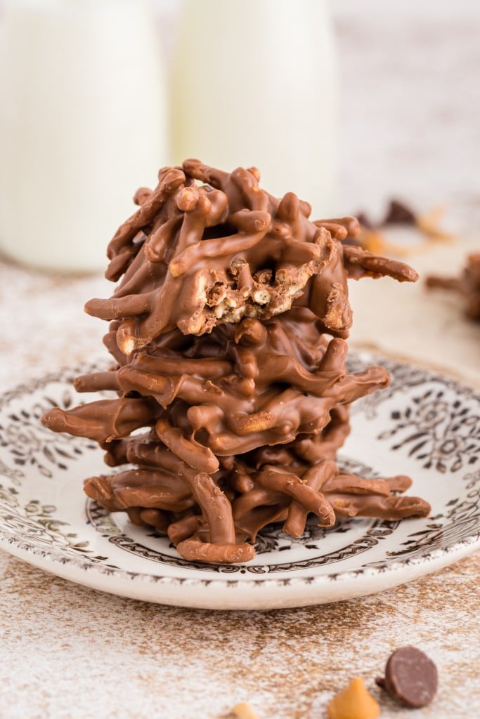 chocolate butterscotch haystacks stacked on a decorative side plate positioned on a destressed wooden surface with two jars of milk in the background