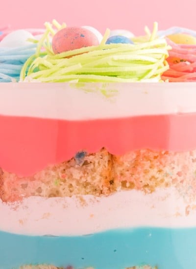 close up of pink, blue, and white layers of pudding and cool whip between squares of funfetti cake, in glass triifle with colored candy "nests and eggs" on top