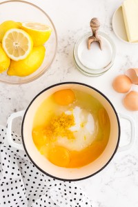 Medium saucepan with sugar, lemon juice, eggs, and lemon zest and surrounded by the lemon curd ingredients on a white marble surface with a polka dot cloth to the side