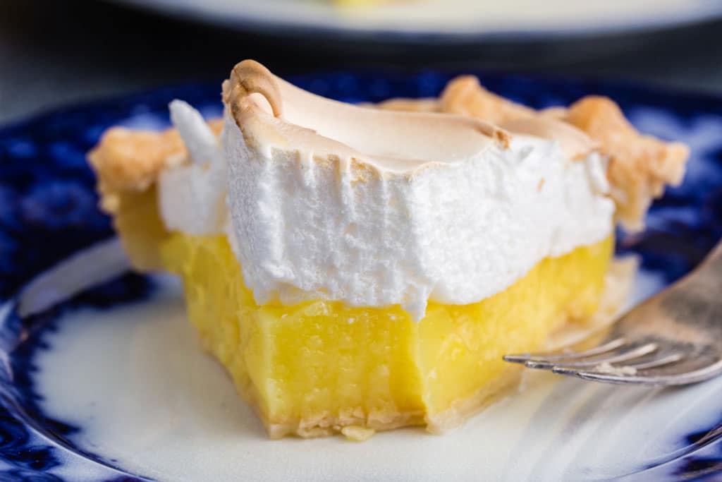 a slice of lemon meringue pie on a blue and white porcelain plate with a serving fork and a piece of pie taken from the tip of the pie slice