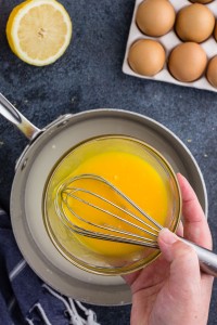 separating eggs yolks from egg whites, pouring the egg yolks into a pan that is placed on a stone-blue surface with cloth and ingredients placed around and whisk ready in hand
