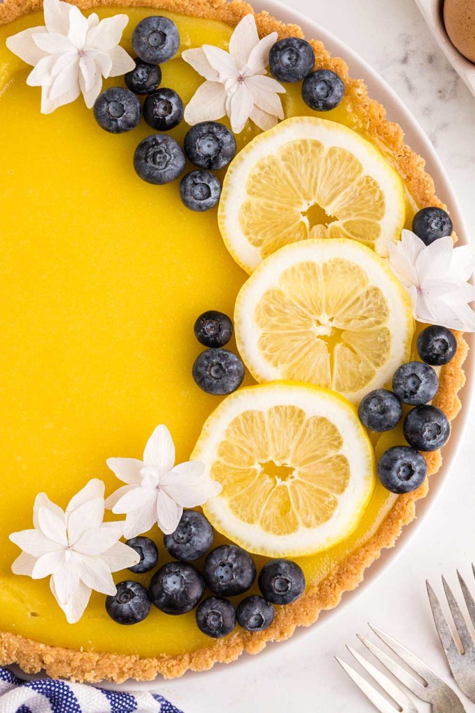Baked and garnished lemon tart with blueberries, slices of lemon and white flowers, seated on a white marble surface with a striped cloth and ingredients positioned around