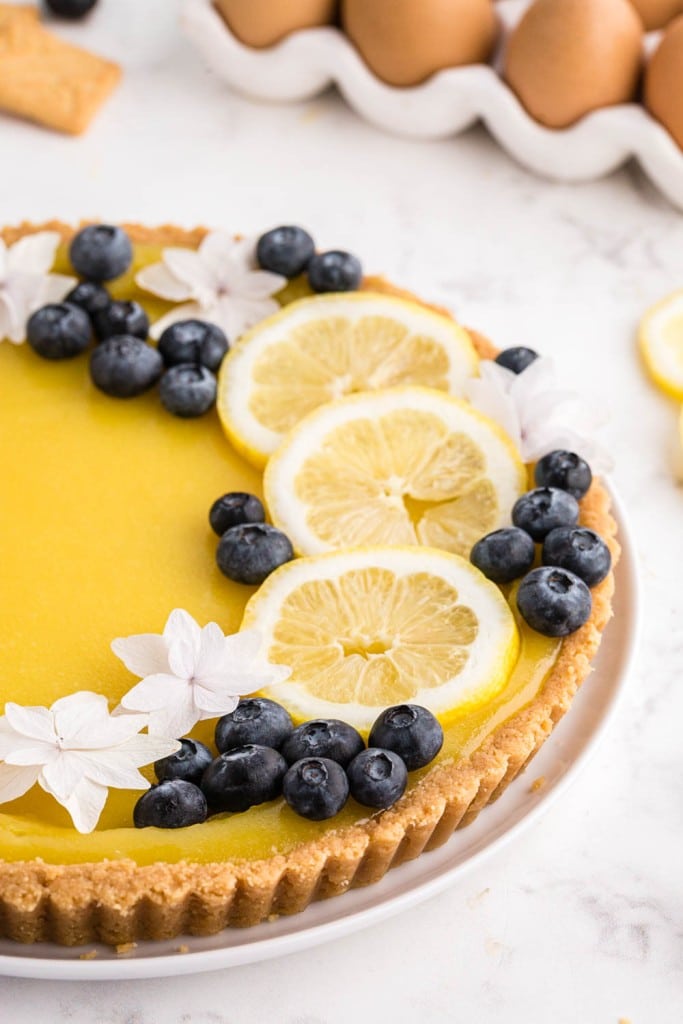 half of a lemon tart captured in the image, garnished with fruit and flowers with ingredients scattered around