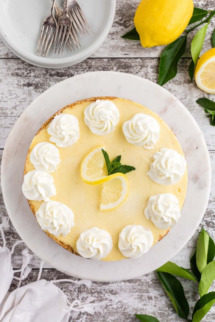 an entire lemon cheesecake viewed from above on a wooden surface with lemons, mint, and a white side plate with cutlery surrounding the cheesecake
