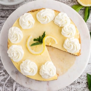 lemon cheesecake viewed from above with a slice removed, garnished with lemon slices and mint and set against a wooden surface
