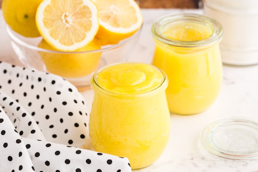 lemon curd placed in two glass jars on a white flat surface with a black and white polka dot kitchen towel and a glass bowl of fresh lemons in the background