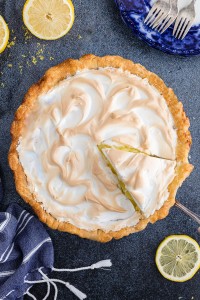 Baked and cooled lemon meringue pie with a slice cut and being removed, lemon slices and cloth placed to the sides of the pie dish and a blue and white serving dish with forks in the corner