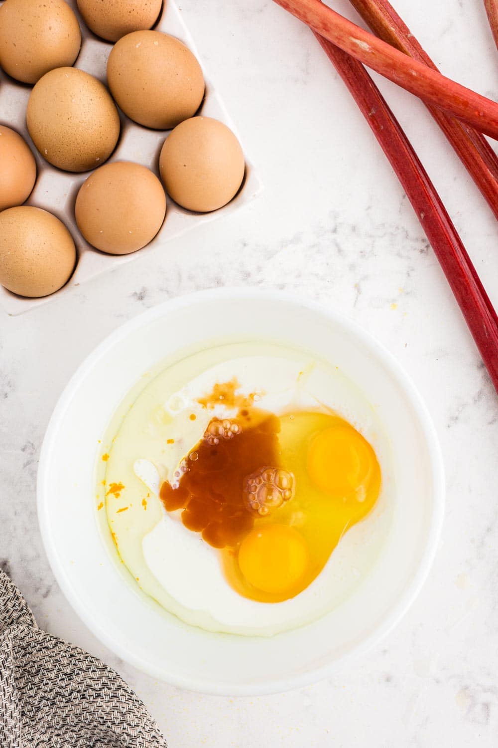 sugar, orange zest, vanilla, vegetable oil, milk, and eggs in a white mixing bowl atop a white marble surface with fresh eggs and rhubarb in the background