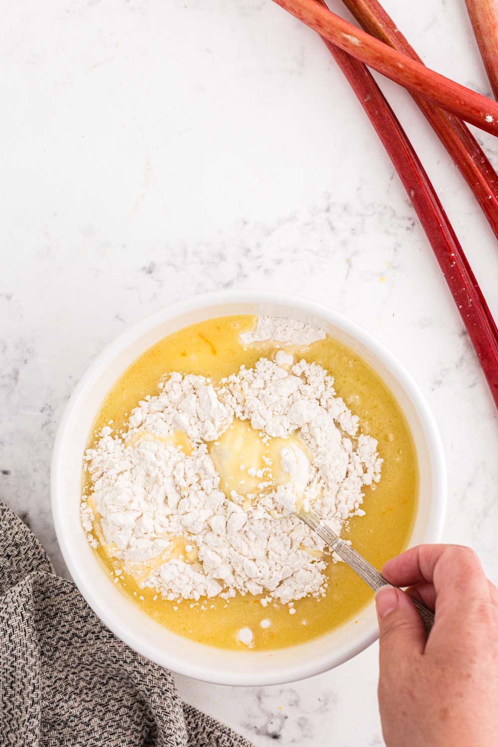 Gently folding the dry ingredients into the wet ingredients in a mixing bowl atop a white marble surface with rhubarb stalks and a kitchen cloth in the background