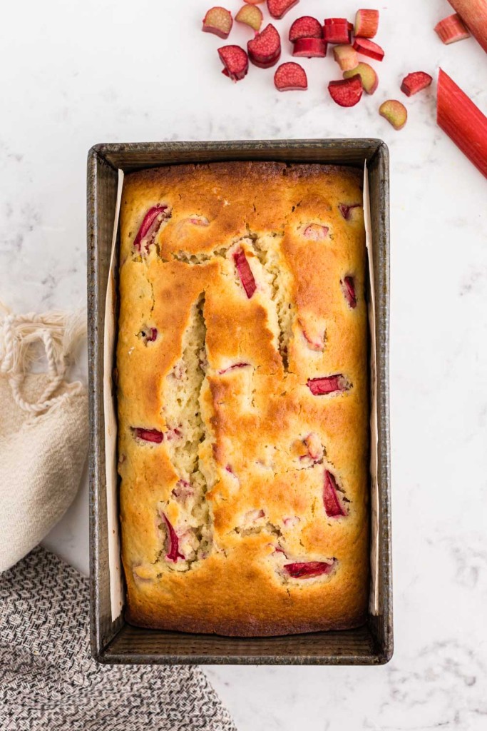 Loaf of baked rhubarb bread in a baking pan set upon a white marble surface with a kitchen towel to the left bottom corner and sliced rhubarb stalks in the right top corner