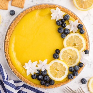 Baked and garnished lemon tart with blueberries, slices of lemon and white flowers, seated on a white marble surface with a striped cloth and ingredients positioned around
