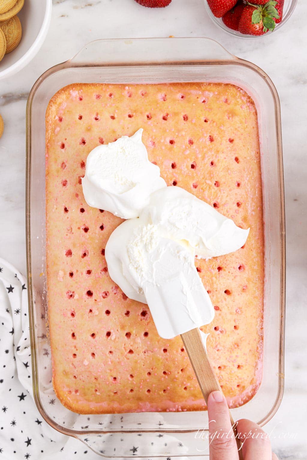 Cool Whip spread over the poke cake atop a white marble surface with Oreos and strawberries in the top corners and a polka dot cloth in the left bottom corner