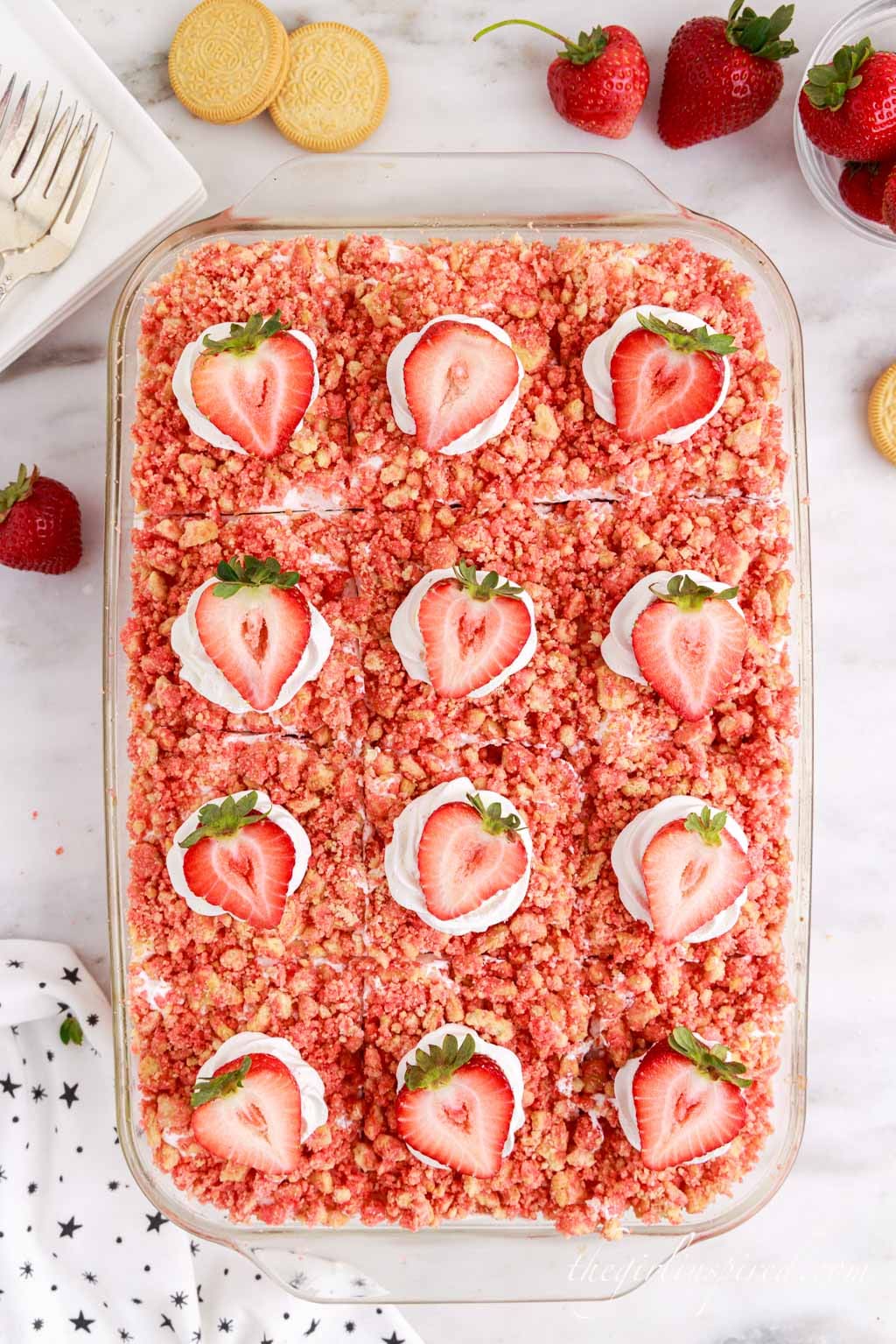 Assembled strawberry crunch poke cake garnished with Cool Whip and halved strawberries, atop a white marble surface with ingredients and a cloth scattered around in the background