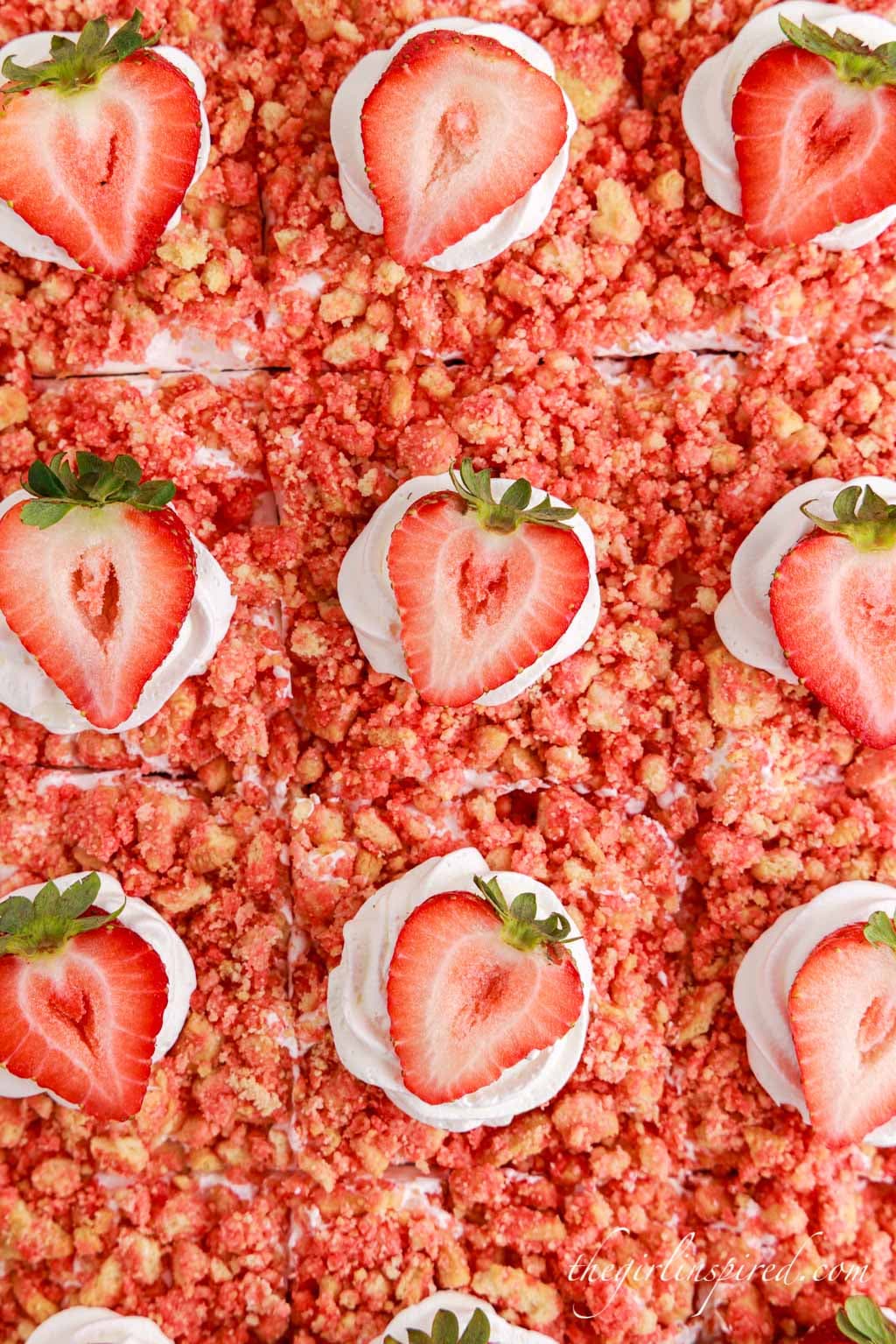 Assembled strawberry crunch poke cake garnished with Cool Whip and halved strawberries, atop a white marble surface with ingredients and a cloth scattered around in the background