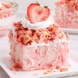 Slice of strawberry crunch poke cake on a white plate atop a white marble surface with additional cake in the background