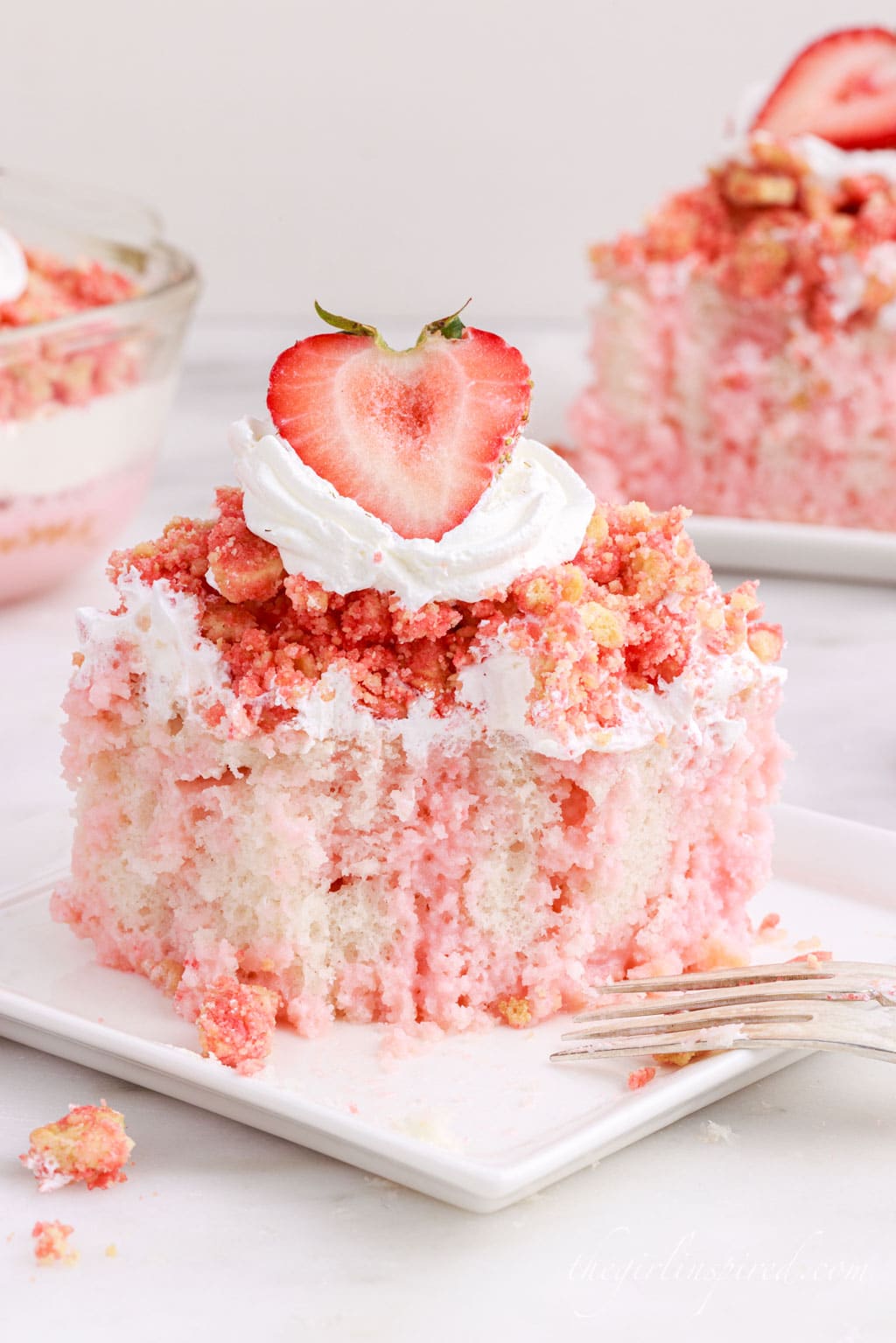 Slice of strawberry crunch poke cake on a white plate with fork atop a white marble surface with extra cake in the background