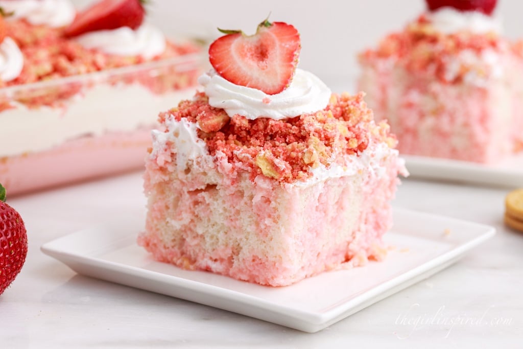 Strawberry crunch poke cake on a white plate atop a white marble surface with additional cake in the background