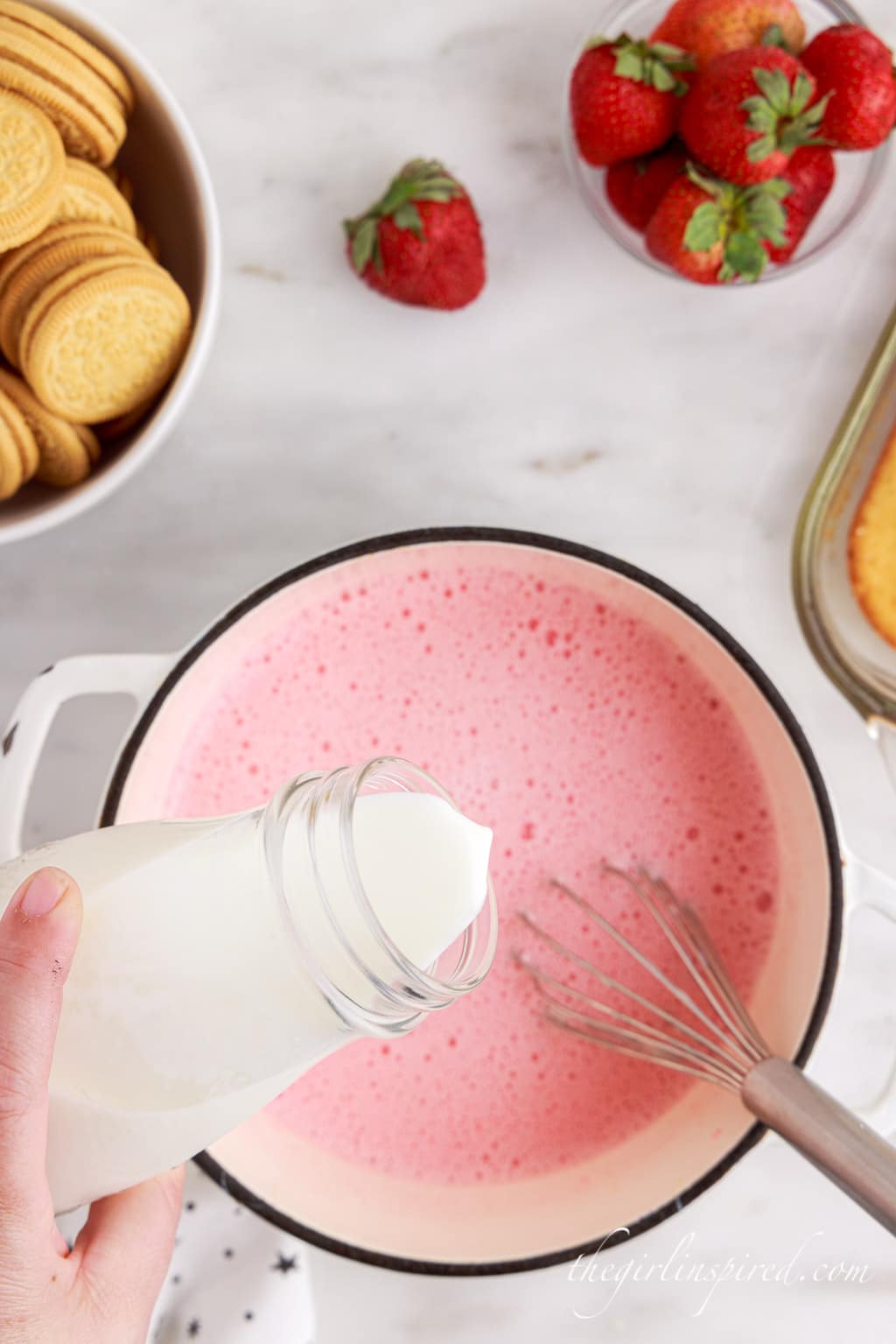 Milk and condensed milk added to the evaporated milk and strawberry gelatin mixture in a saucepan atop a white marble surface with Oreos and fresh strawberries in the background