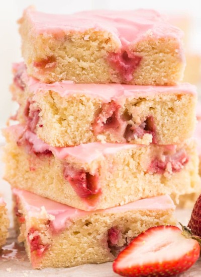four strawberry lemon blondies piled on top of each other atop a white marble surface with sliced strawberry in the front