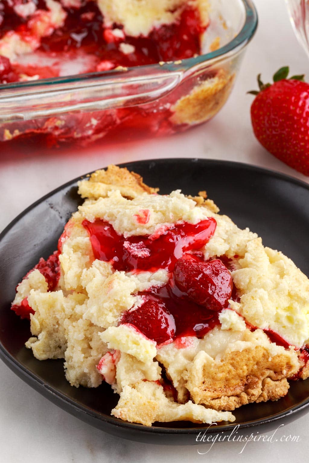 A serving of strawberry cheesecake dump cake on a black plate atop a white marble surface with baking dish and cloth in the background.
