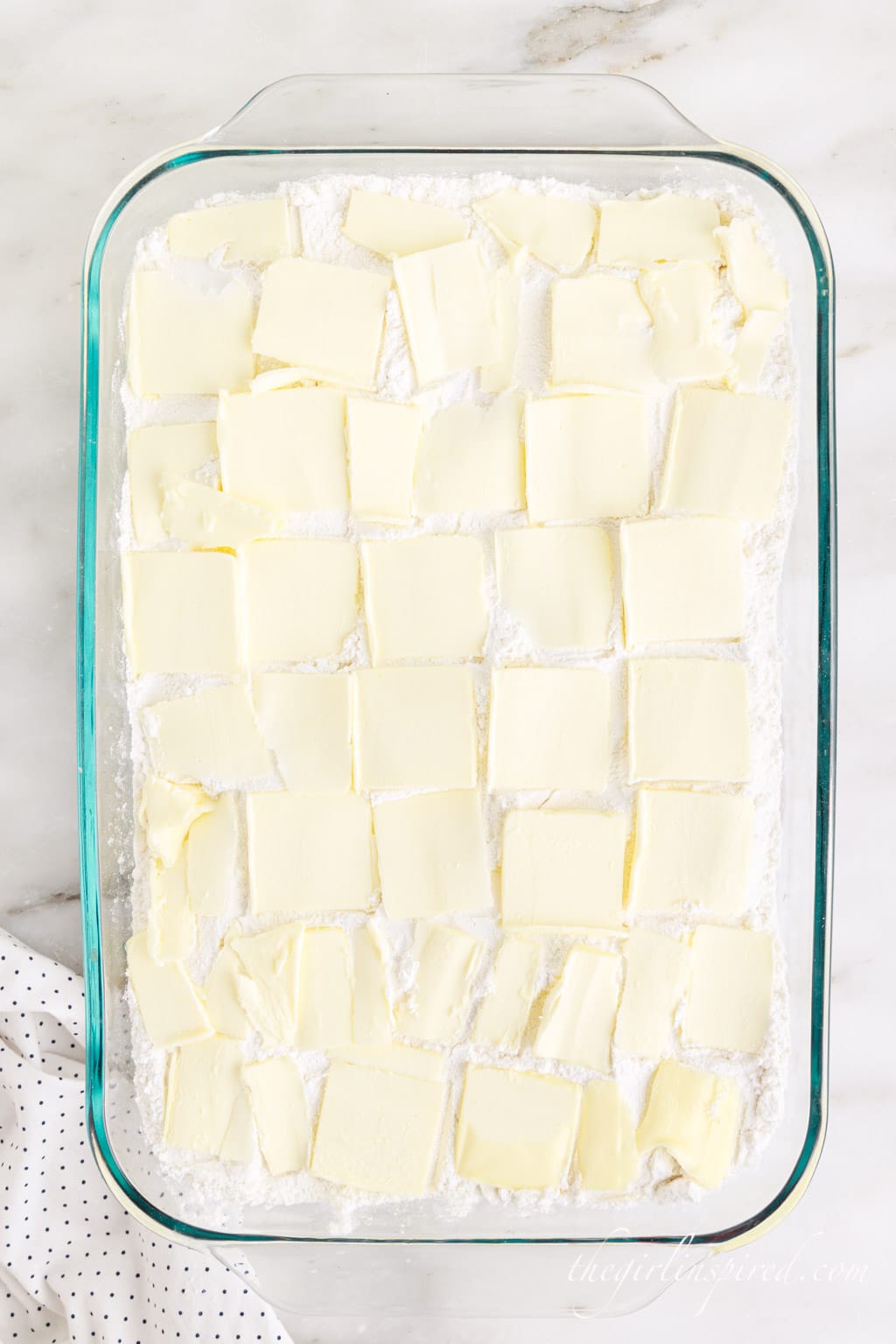 Thin pats of butter added to the top of the dump cake layers in the glass baking dish atop a white marble surface with a white cloth in the bottom left corner