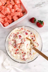 Fresh strawberries added to the cheesecake mixture with cubed cake and strawberries in the background, atop a white marble surface, cloth in the bottom left corner
