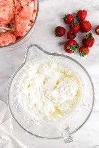 Heavy cream whipped in a glass bowl, atop white marble surface with cubed cake and strawberries in the background