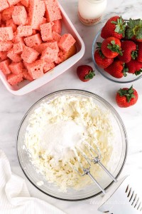 Bowl of cream cheese and extract mixed with an electric mixer with cubed cake and fresh strawberries in the background, cloth in the bottom left corner