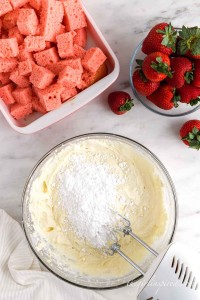 Powdered sugar added to the cream cheese and pudding mixture with cubed cake and fresh strawberries in background, atop a white marble surface, cloth in the bottom left corner