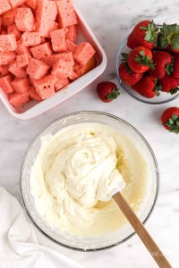 Cheesecake instant pudding mixed into the cream cheese mixture with cubed cakes and fresh strawberries in background, atop white marble surface, cloth in the left bottom corner