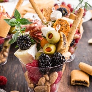 Clear plastic cup with crackers, berries, brie wedge, olives, breadsticks, and nuts.