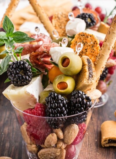 Clear plastic cup with crackers, berries, brie wedge, olives, breadsticks, and nuts.