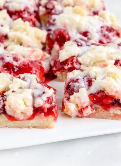 Cherry pie bars plated on a white platter with cherry pie mixture in a glass bowl, forks, and white plates in the background