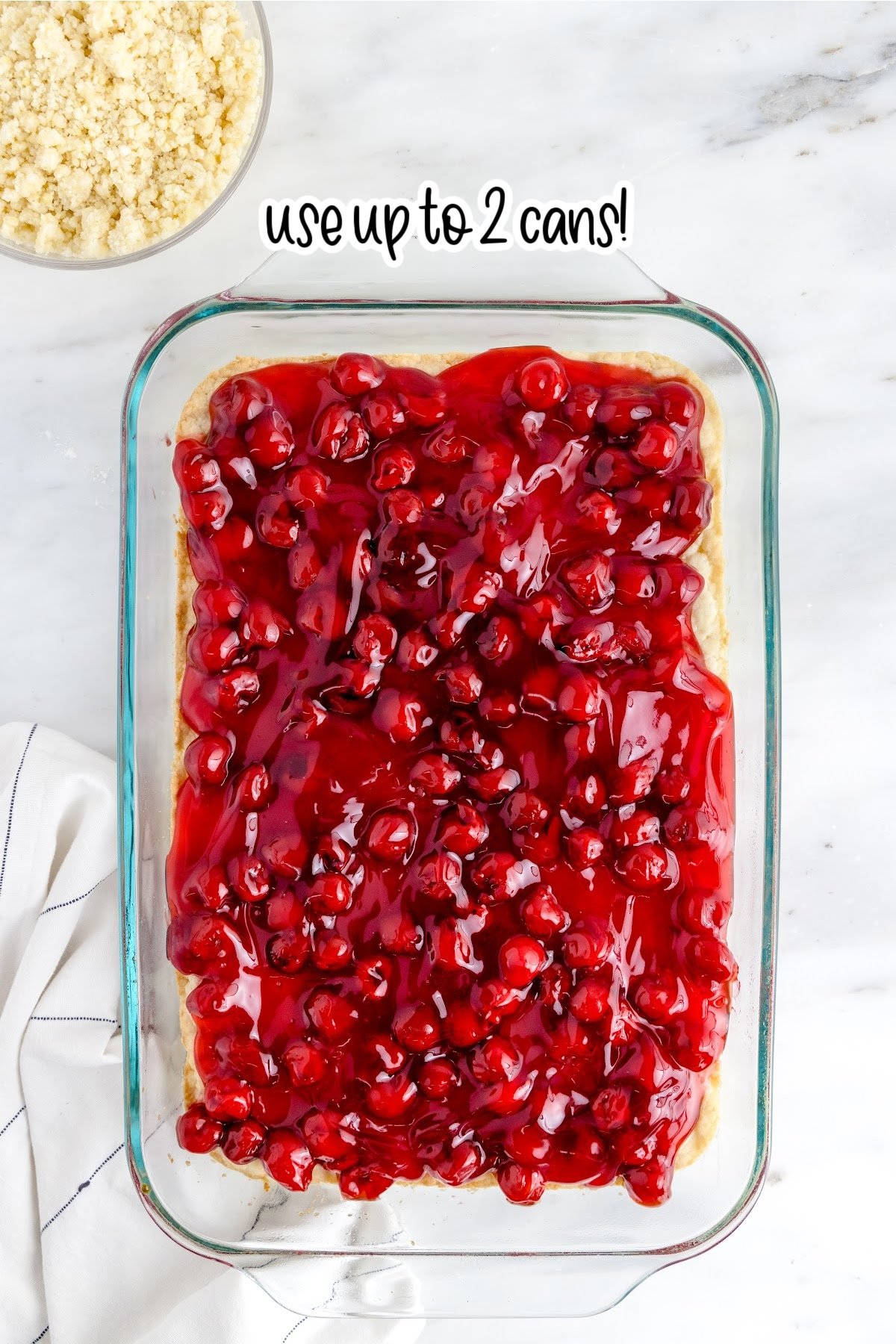 cherry pie filling spread over the baked dough crust on top of a white marble countertop with a cloth next to the baking dish and text "use up to 2 cans."