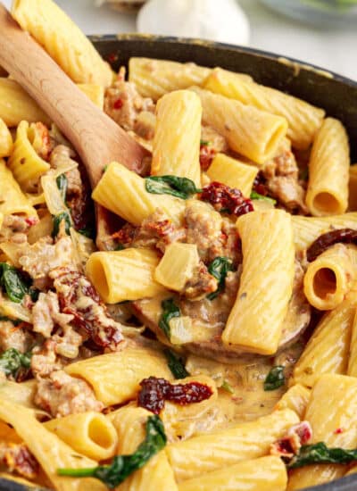 Wooden spoon scooping Italian sausage pasta with big chunks of pasta, ground sausage, sun-dried tomatoes, and wilted spinach from a creamy sauce in skillet.