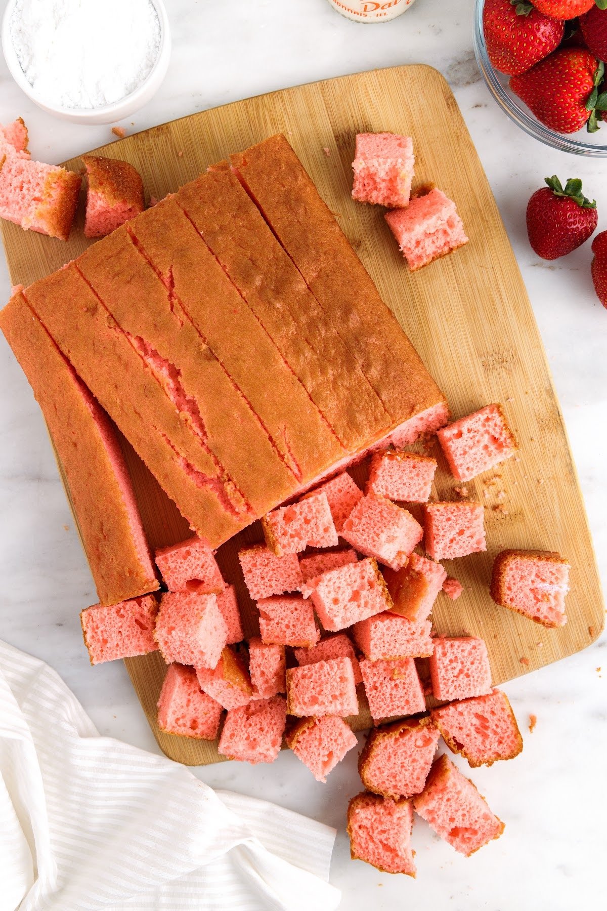 Strawberry sheet cake on cutting board, some sliced into cubes of cake.
