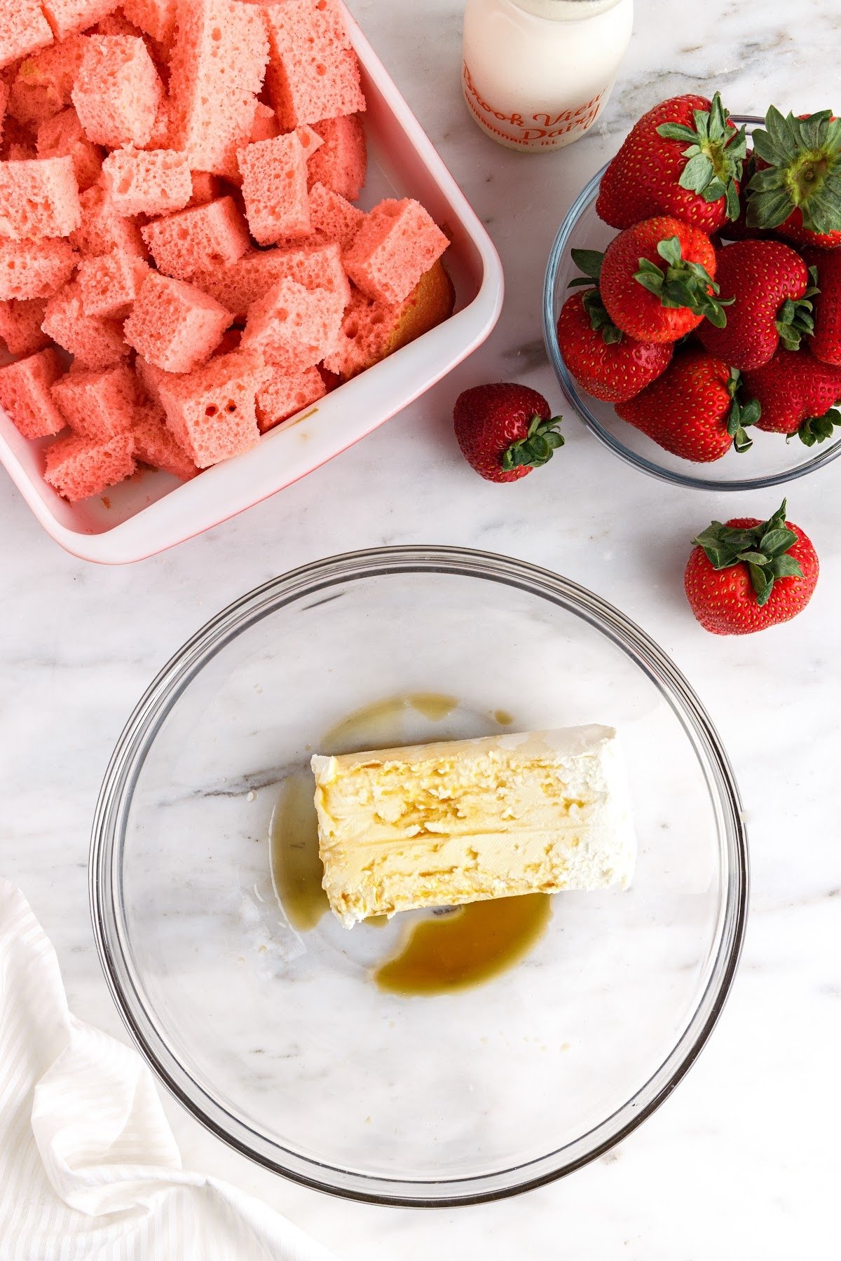 Block of cream cheese and vanilla extract in glass bowl, next to cubes of strawberry cake and bowl of fresh strawberries.
