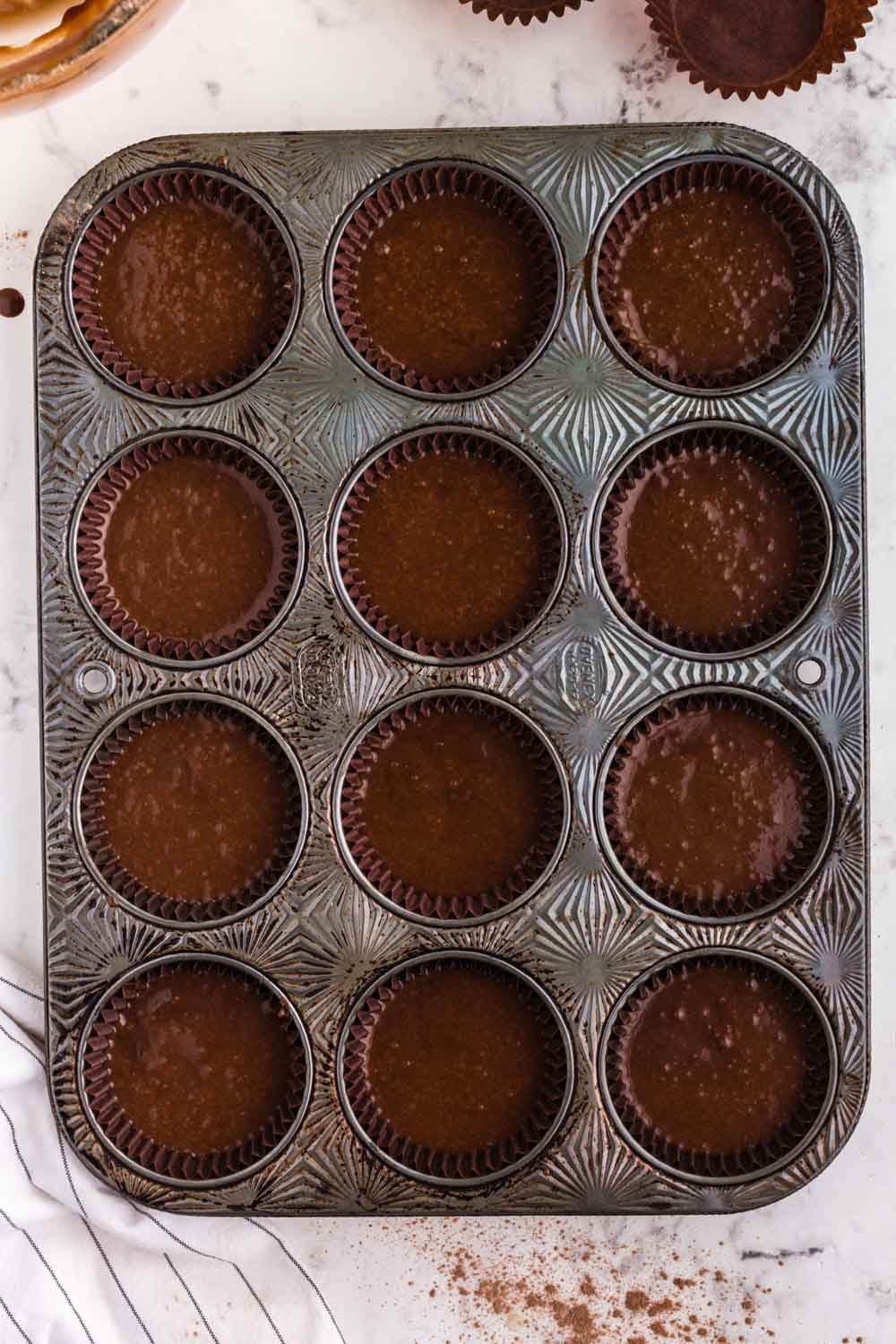 Chocolate cupcake batter poured into prepared metal muffin tin, on top of a marble surface, linen cloth in background