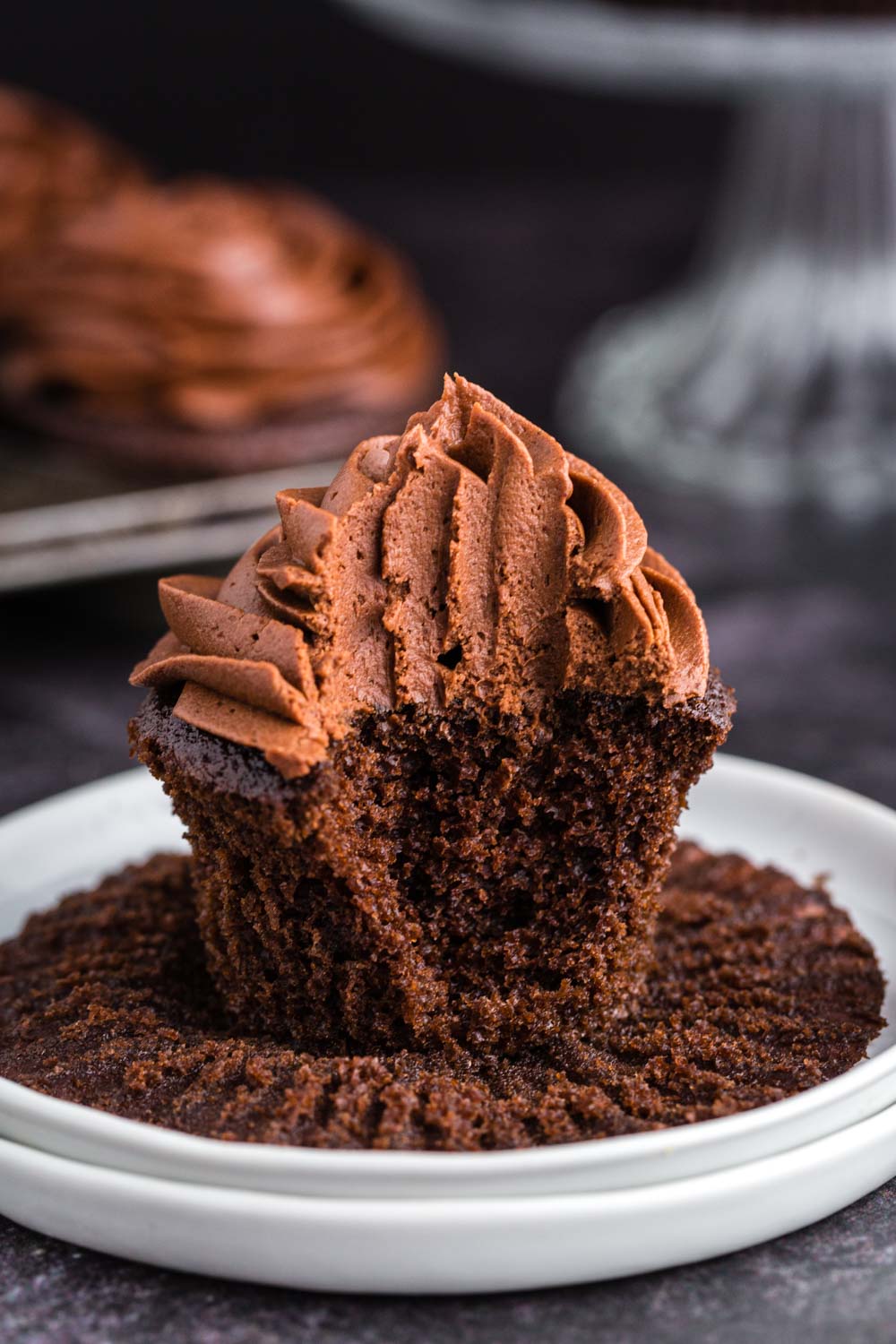 half eaten chocolate cupcake with chocolate buttercream frosting, unwrapped cupcake liner, on top of stacked white side plates with frosted cupcakes in a tin pan in background