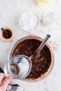 Boiling water added to the chocolate cake batter in a glass mixing bowl, extra batter ingredients in separate containers, black and white checked cloth, on top a white marble surface