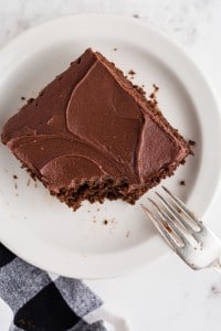 Single slice of classic chocolate cake served on a white plate with a fork, black and white checked cloth, on top of a white marble surface