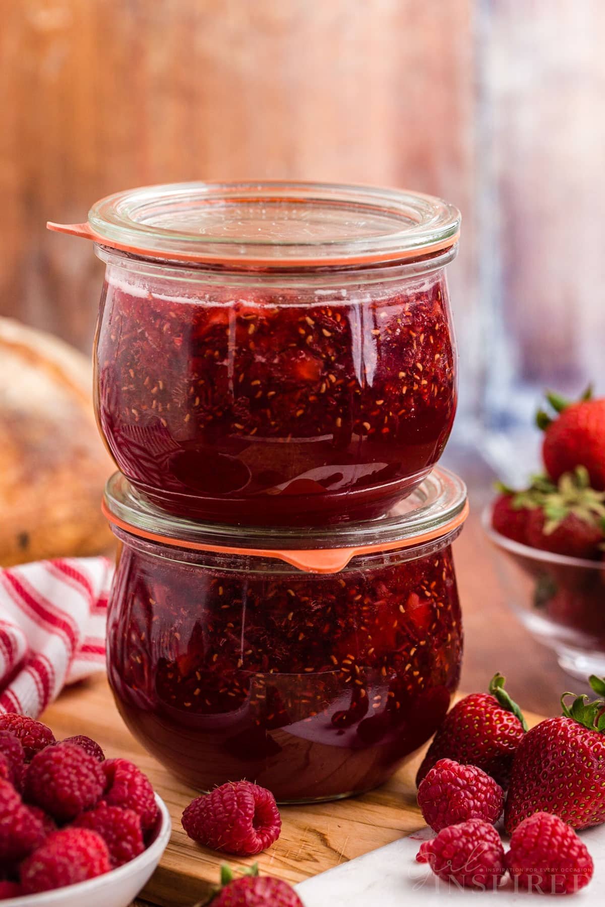 Two stacked glass jars filled with slow cooker strawberry and raspberry jam, on top a wooden board, bowl of fresh raspberries and loose raspberries and strawberries on a wooden surface, red and white striped linen cloth