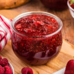 Crockpot Strawberry raspberry jam in glass jar, on top wooden board, bowl of fresh raspberries, bowl of fresh strawberries, red and white striped linen cloth