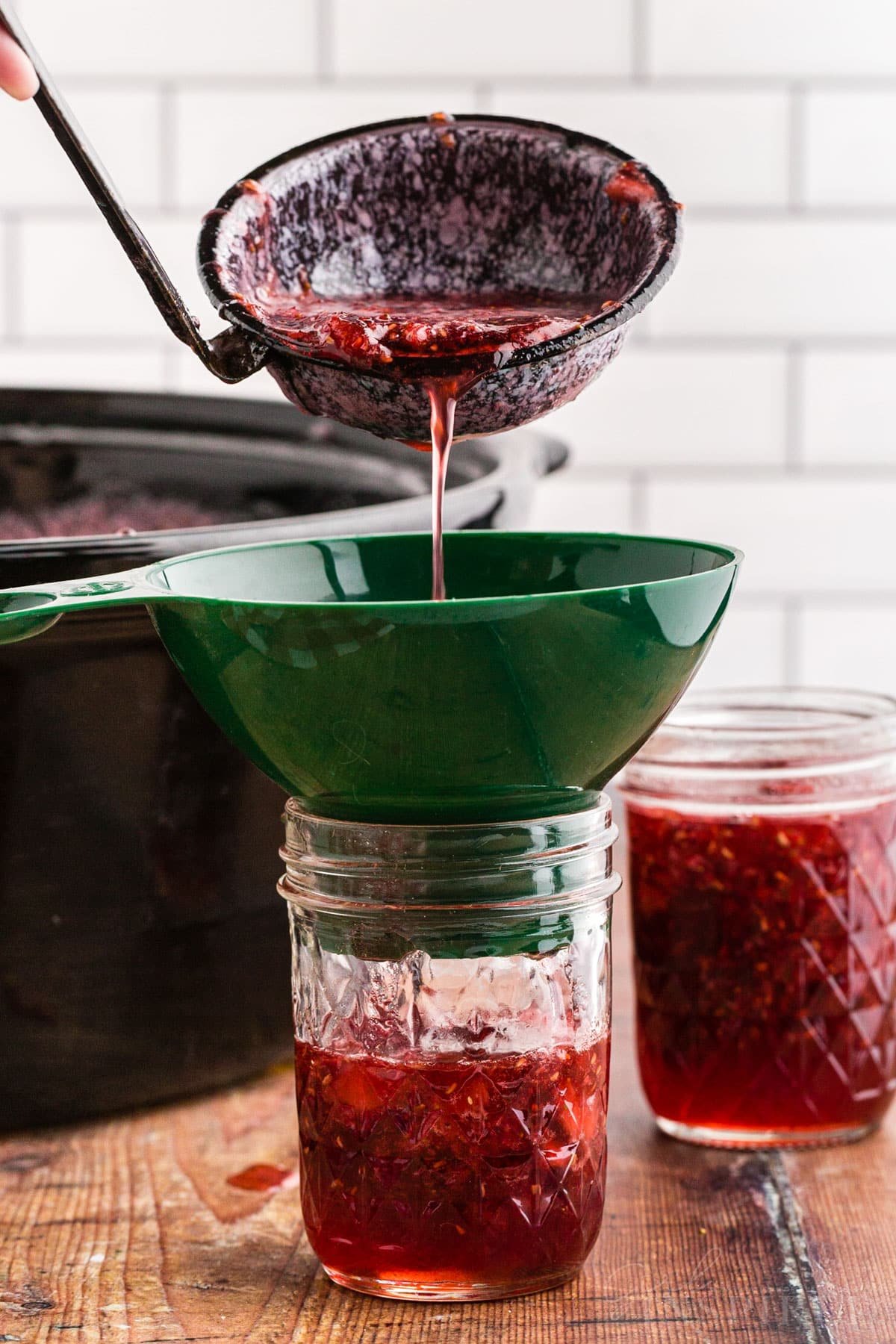 Ladle pouring the strawberry raspberry jam into a jar with the use of a canning funnel placed on top of the jar.