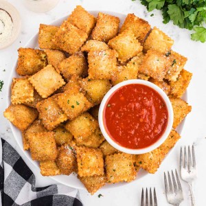 Fried ravioli on a white plate with a bowl of marinara sauce, black and white checked linen, fresh parsley, forks, on a white marble surface
