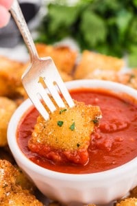 Fried ravioli with a bowl of marinara sauce, fresh parsley and black and white checked linen in the background, fork with fried ravioli dipped into sauce