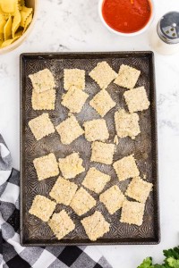 baking sheet with coated breadcrumbs, bowl of refrigerated ravioli, bowl of marinara sauce, black and white checked linen, fresh parsley, on a marble countertop