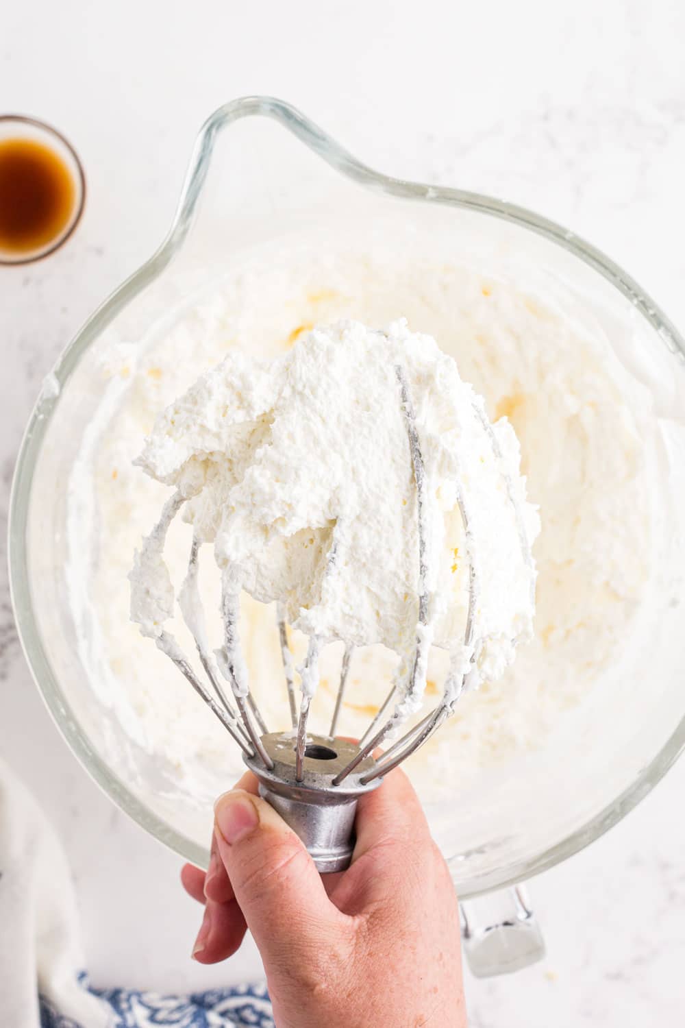Metal whisk attachment above bowl of whisked egg whites, sugar mixture, cubed butter, and cream of tartar, bowl of vanilla extract, blue and white decorative linen, on a marble countertop